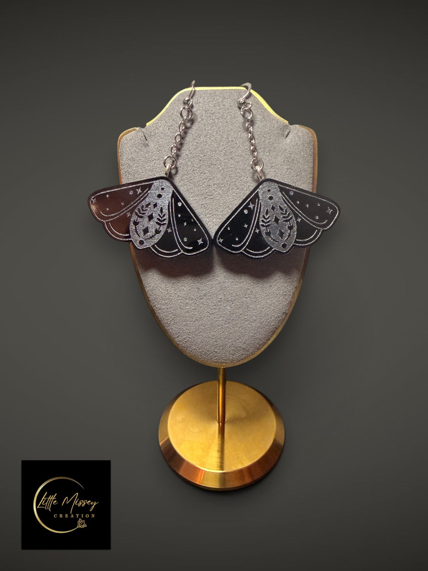 Little Missey Creation black resin moth dangle earrings with celestial silver engravings on a grey display stand