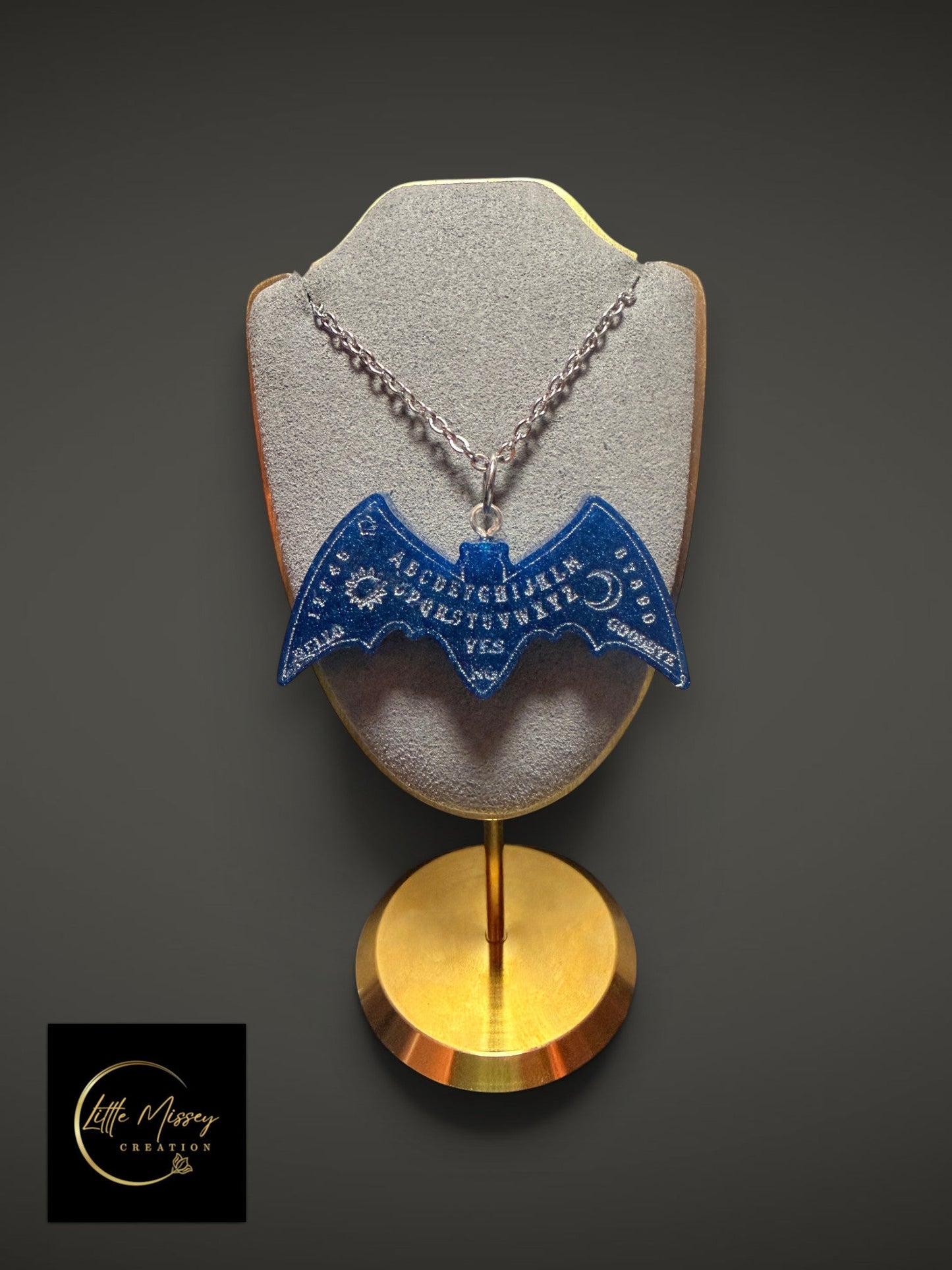Little Missey Creation bat-shaped Ouija board pendant necklace in glittery blue resin on a grey display stand.