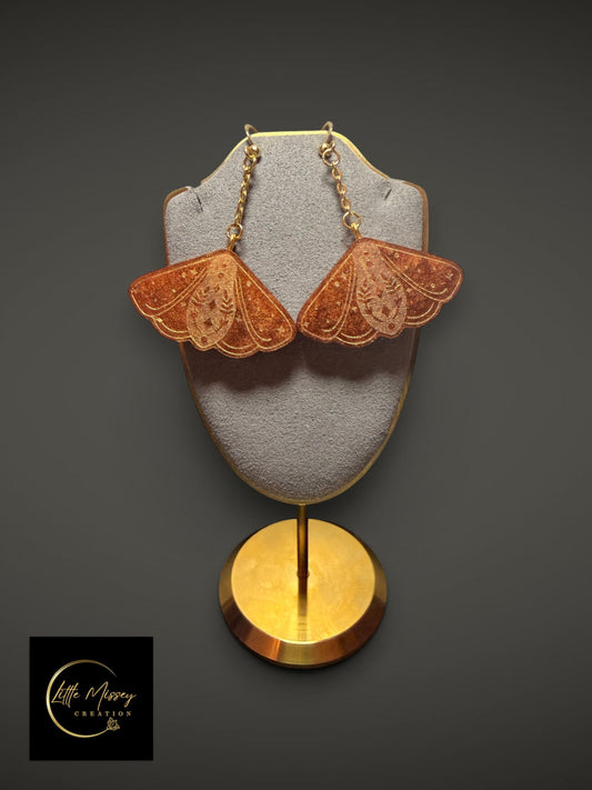 Little Missey Creation golden glitter resin moth dangle earrings with celestial engravings on a grey display stand